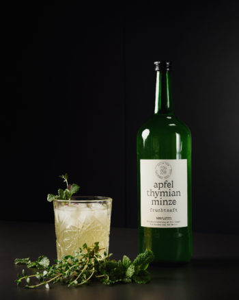 Flasche Apfel-Thymian-Minze-Fruchtsaft von wwurm neben einem Glas mit Eis und frischen Kräutern, vor dunklem Hintergrund